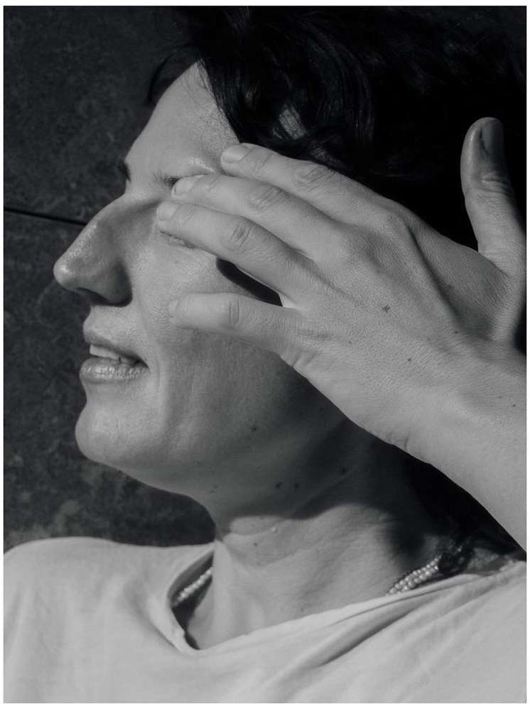 Monochrome portrait of an adult woman with dark hair, hand shielding her eyes, wearing a light shirt and necklace, outdoors.