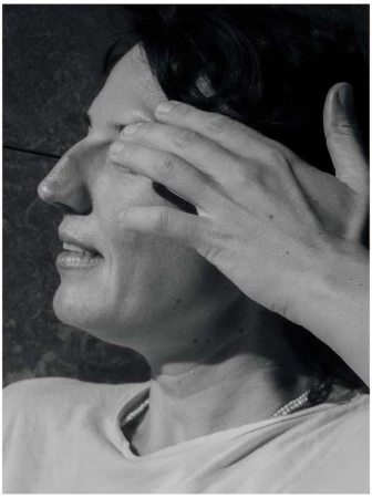 Monochrome portrait of an adult woman with dark hair, hand shielding her eyes, wearing a light shirt and necklace, outdoors.