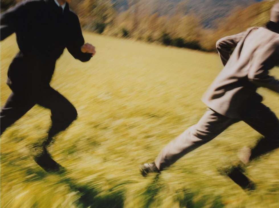 Motion-blurred photo of two people running across a grassy field; one in a dark suit and the other in a light suit. Landscape background.