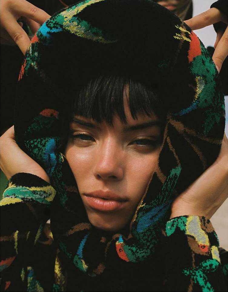 Close-up of a young woman with dark hair framed by a patterned fabric wrap held by hands, set against a neutral background.