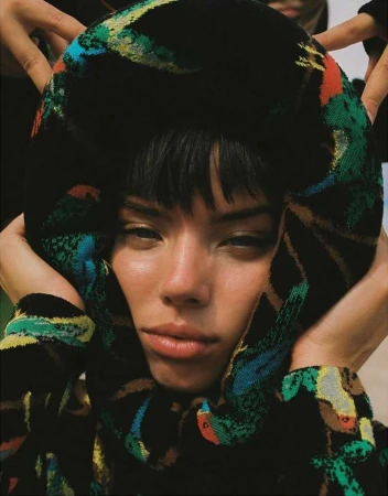 Close-up of a young woman with dark hair framed by a patterned fabric wrap held by hands, set against a neutral background.