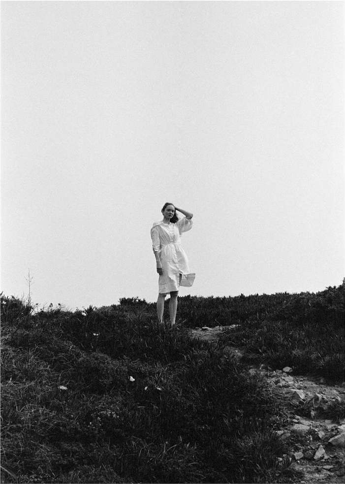 Woman in a white dress standing on a grassy hill under an overcast sky, captured in monochrome by photographer Iris Humm.