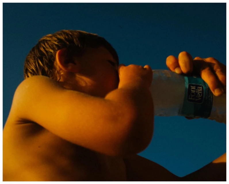 A young child with fair skin is drinking from a clear plastic water bottle held by an adult's hand against a bright blue sky background.