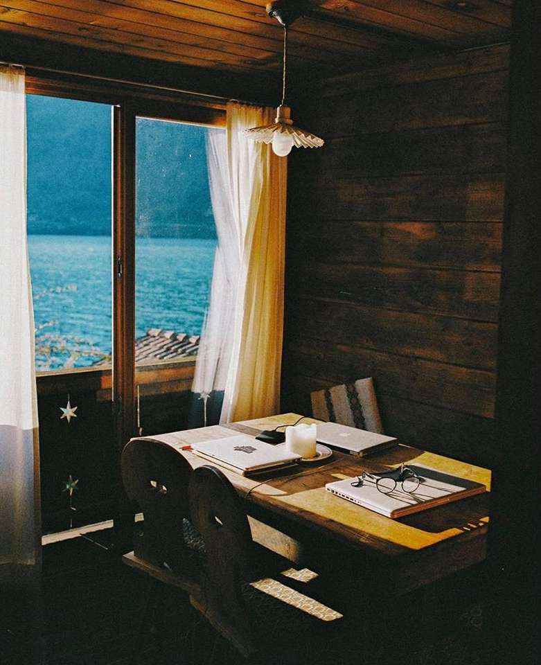 Indoor view of wooden table with laptops, eyeglasses, and a candle, framed by a window showing a blue water landscape.