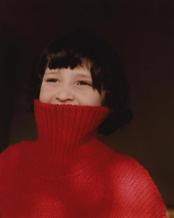 Young girl with short dark hair smiles in a red knit turtleneck sweater against a dark background in a medium indoor studio shot.