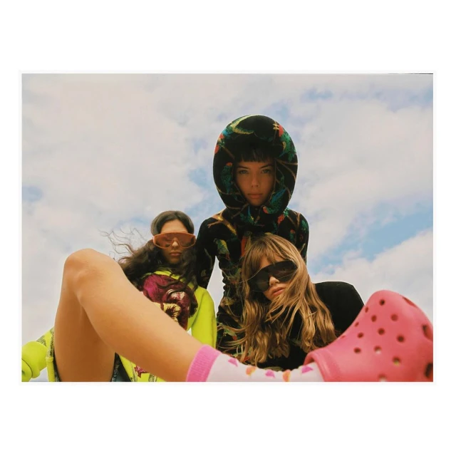 Three young women pose in colorful, patterned hoodies and sunglasses with a pink clog in foreground against a blue sky backdrop.