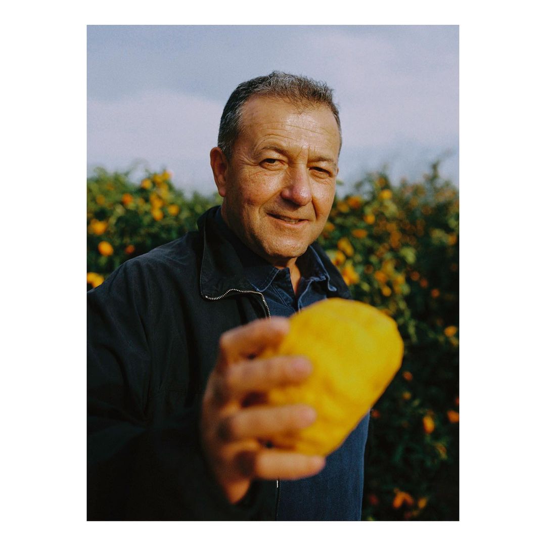 An adult man holding a large, yellow etrog fruit outdoors during the day, with a backdrop of trees and a cloudy sky.
