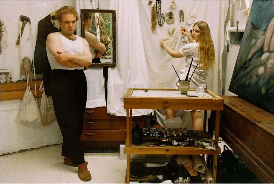 Interior of an artist's studio with two young adults, one male, one female, surrounded by art supplies, paintings, and draped white fabric.