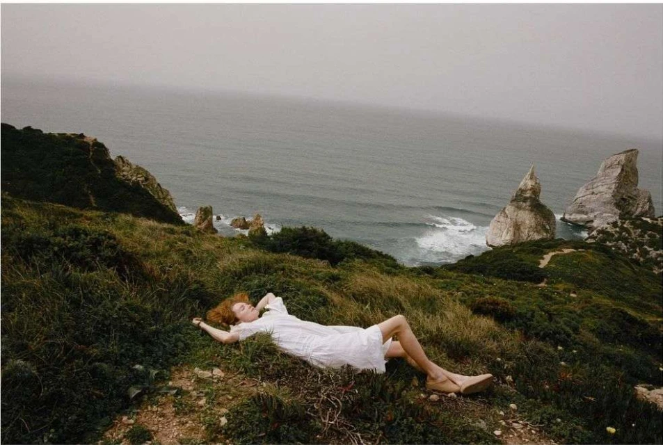 A woman in a white dress lies on a grassy cliff overlooking the ocean with rock formations. Overcast sky. Relaxation theme.