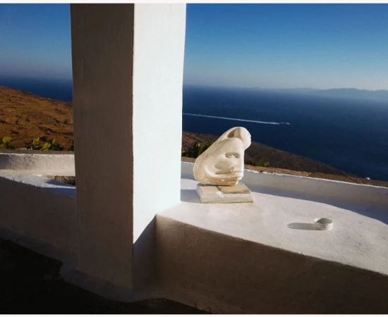 A stone head sculpture sits on a white ledge overlooking a blue ocean and hills, viewed through a white archway on a sunny day.