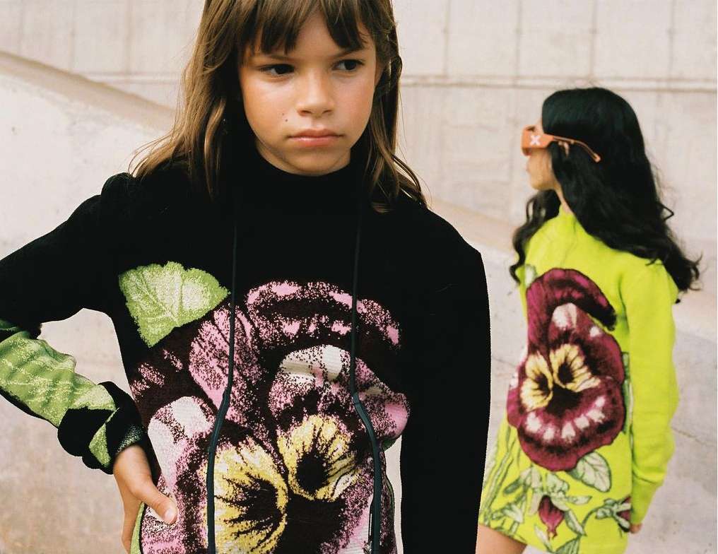 Two young children pose outdoors wearing floral print apparel, one in black and one in green, against a neutral background.