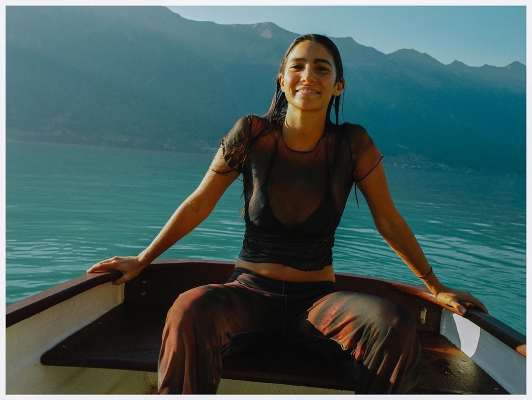Woman in boat on a lake with mountains in background. She wears dark top and patterned trousers, and smiles in outdoor setting.