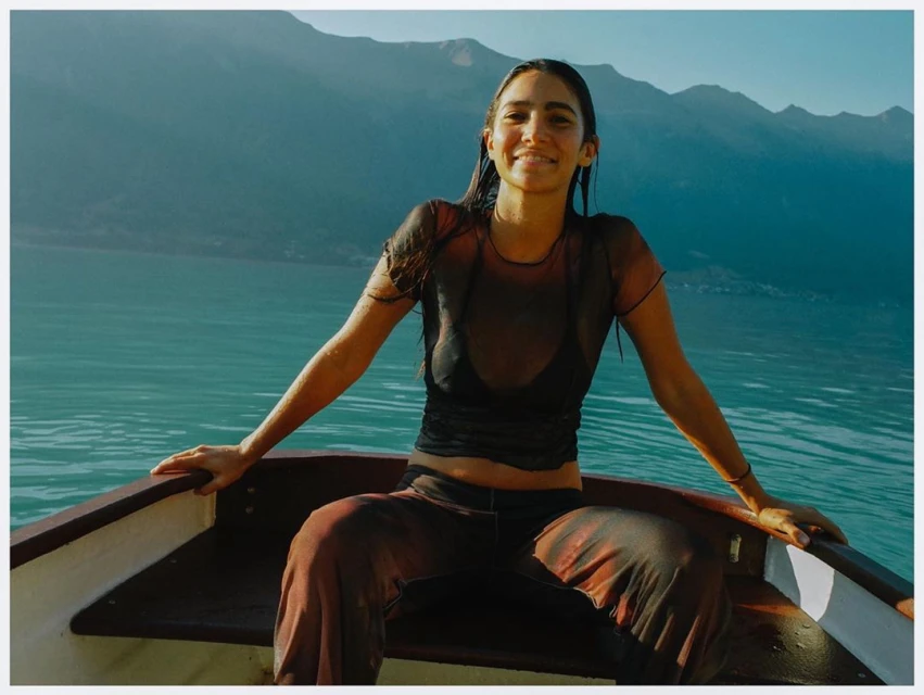 Woman in boat on a lake with mountains in background. She wears dark top and patterned trousers, and smiles in outdoor setting.