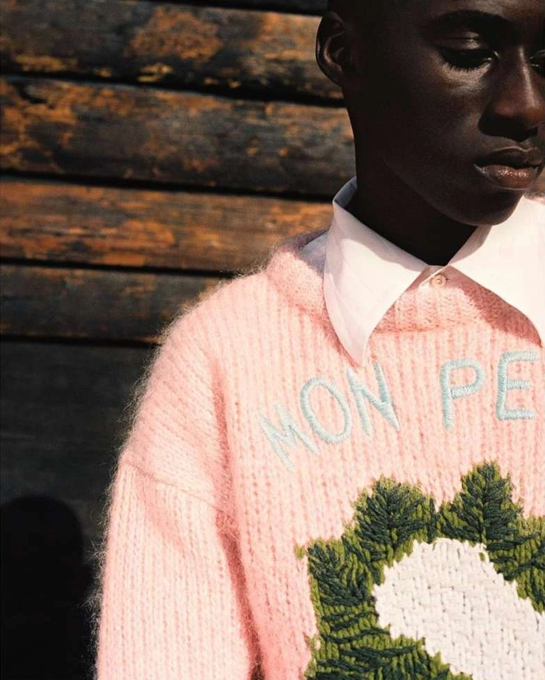 Close-up portrait of a Black man wearing a fuzzy pink knitted sweater and a white collared shirt against a dark wooden wall.