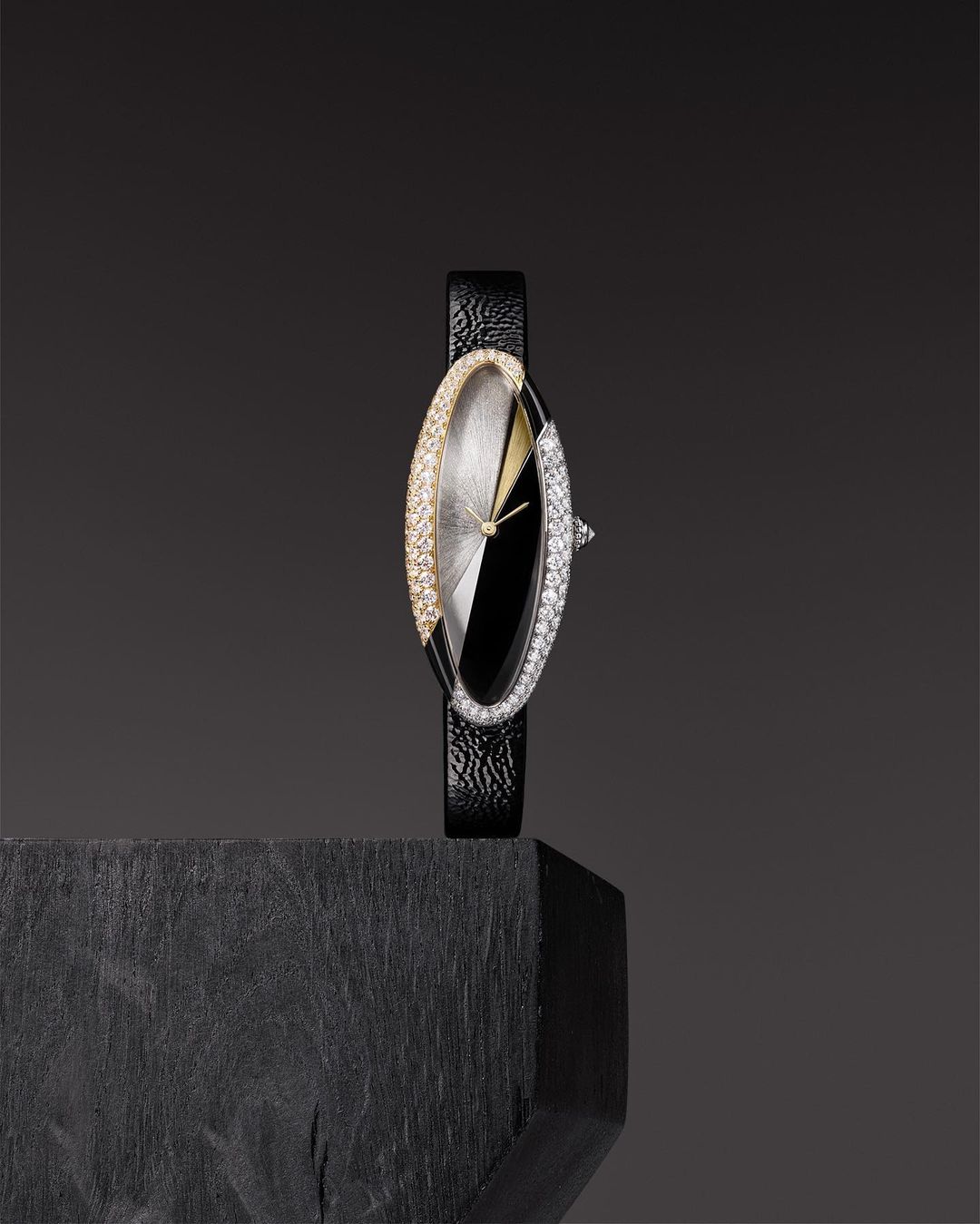 Elegant wrist watch with an oval diamond-encrusted face against a dark gray background, resting on a black textured surface.