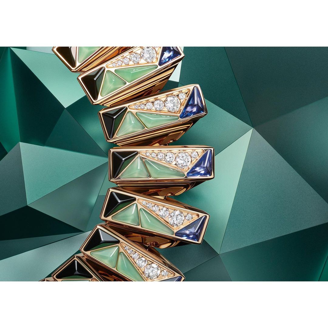 Close-up of a geometric gold ring with green, blue, black, and clear gemstones on an emerald green faceted background. Jewelry advertisement.