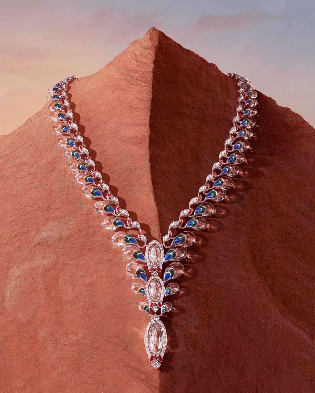 Elegant necklace with diamonds, sapphires, and rubies set against a textured earth tone background evoking a mountain peak and sky.