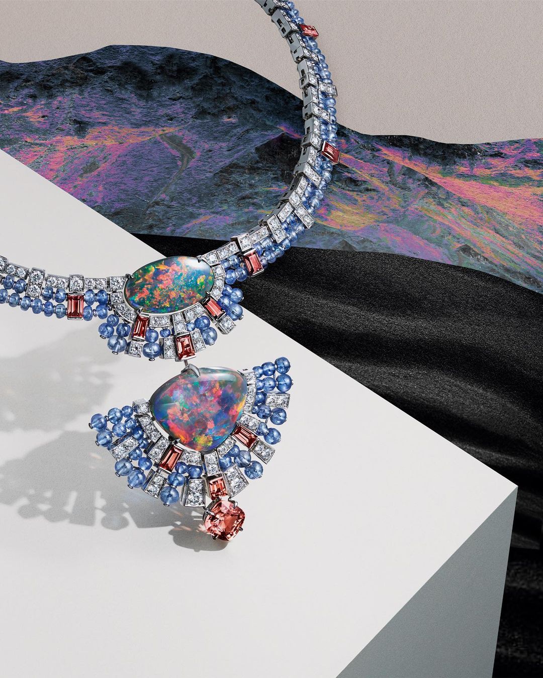 Elegant necklace with opal pendant, set with sapphires and diamonds, displayed against a background featuring both a textured drapery and mountain