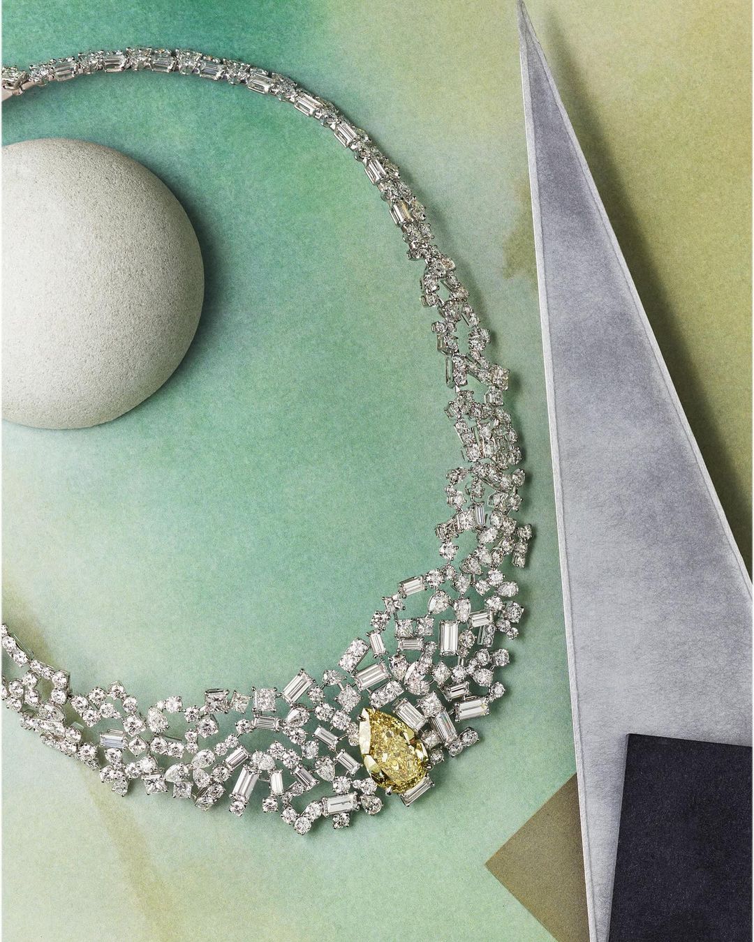 Elegant diamond necklace with a yellow central stone, set on a green watercolor paper background with geometric shapes.