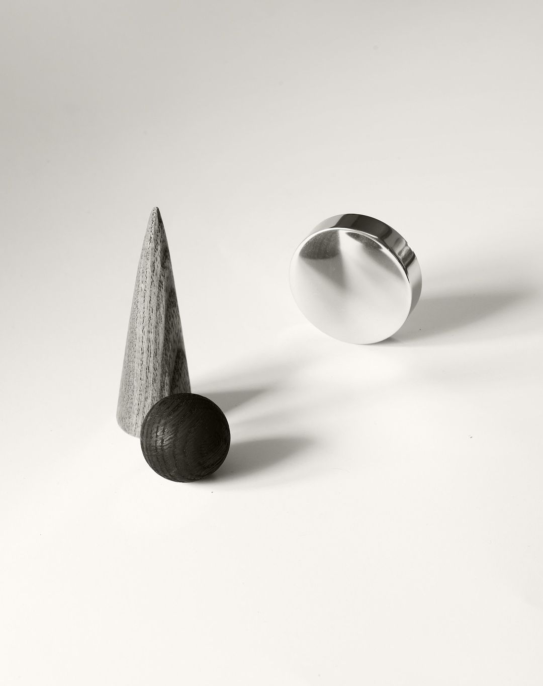 Still life featuring wooden cone, ball, and shiny metal cylinder on a white surface, showcasing minimalist design and geometric shapes.