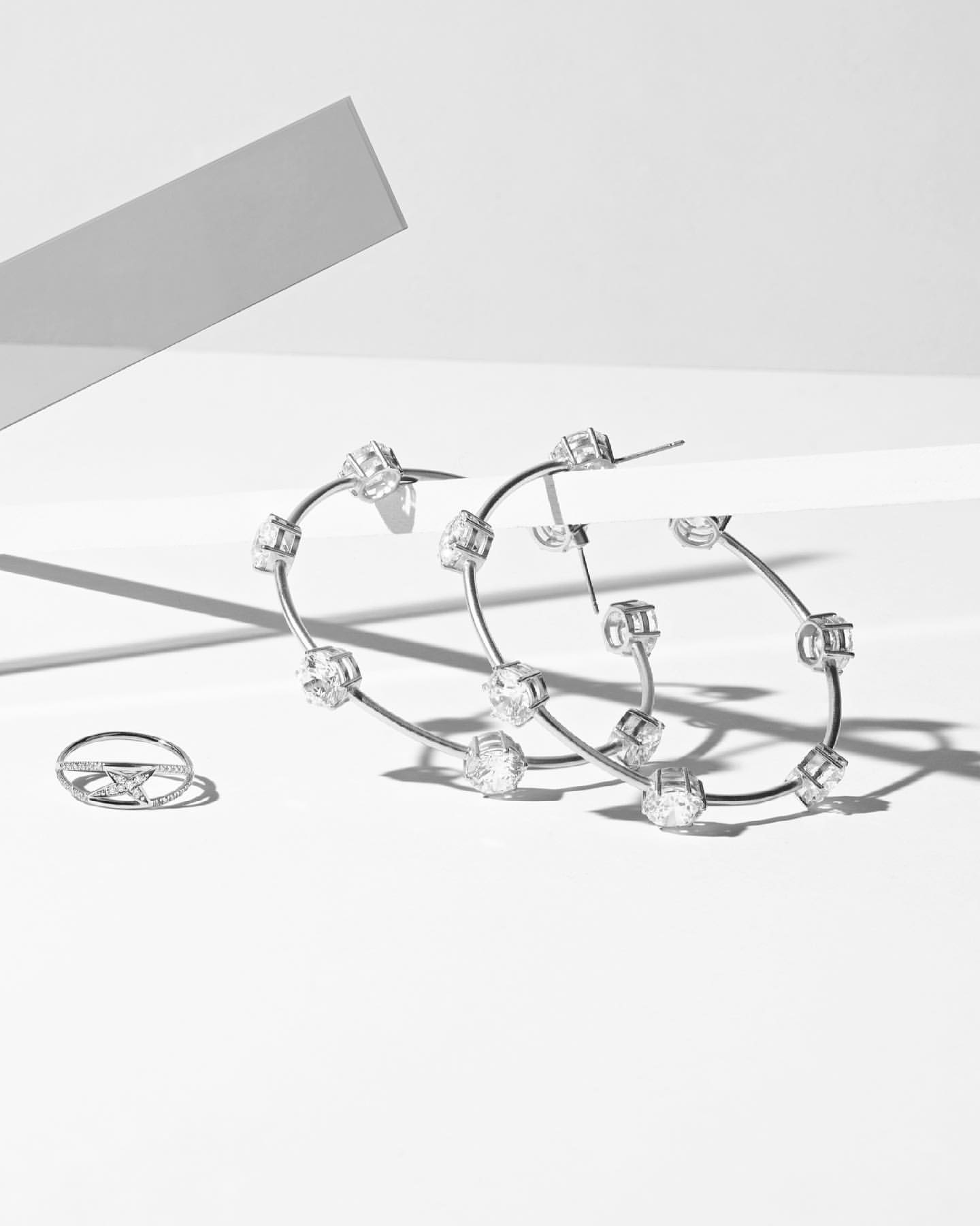 Elegant jewelry still life: Silver hoop earrings with diamond accents and matching ring on a pristine white surface with geometric shadows.