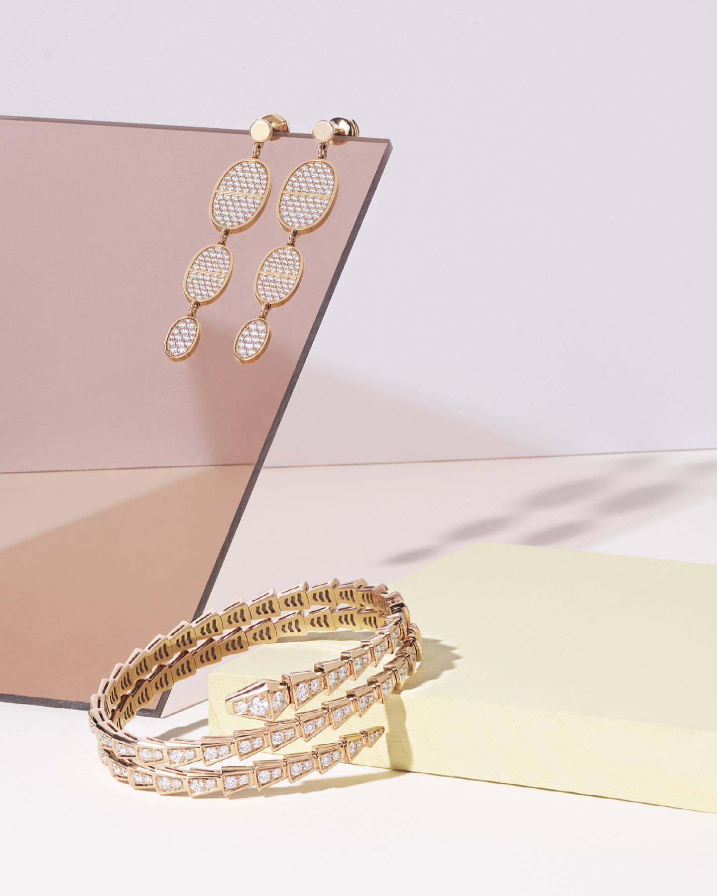 Elegant gold jewelry displayed against pastel geometric shapes, featuring a coiled bracelet and long, drop earrings, studio lit.