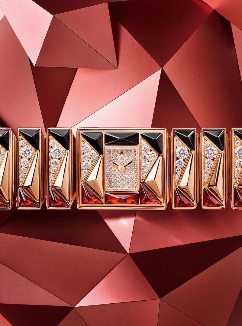 Close-up of a geometric watch with gold, black, and gemstone accents. The watch face features diamond pavé on a triangular background.