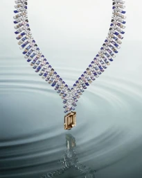 An elaborate diamond and gemstone necklace suspended above rippling water with a clear reflection.