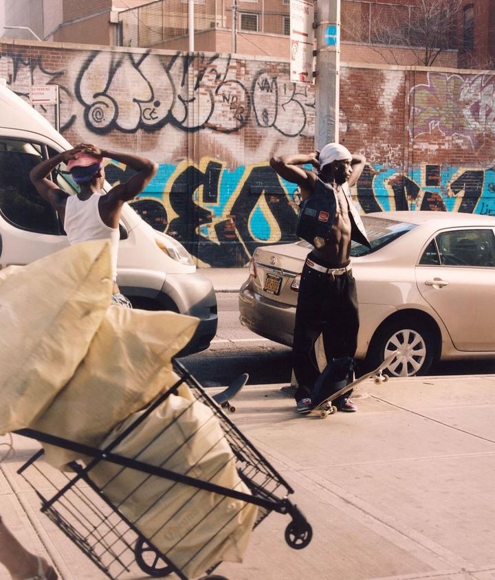 Two men pose in an urban setting next to cars and a brick wall covered in graffiti, one with a skateboard, shopping cart in foreground.