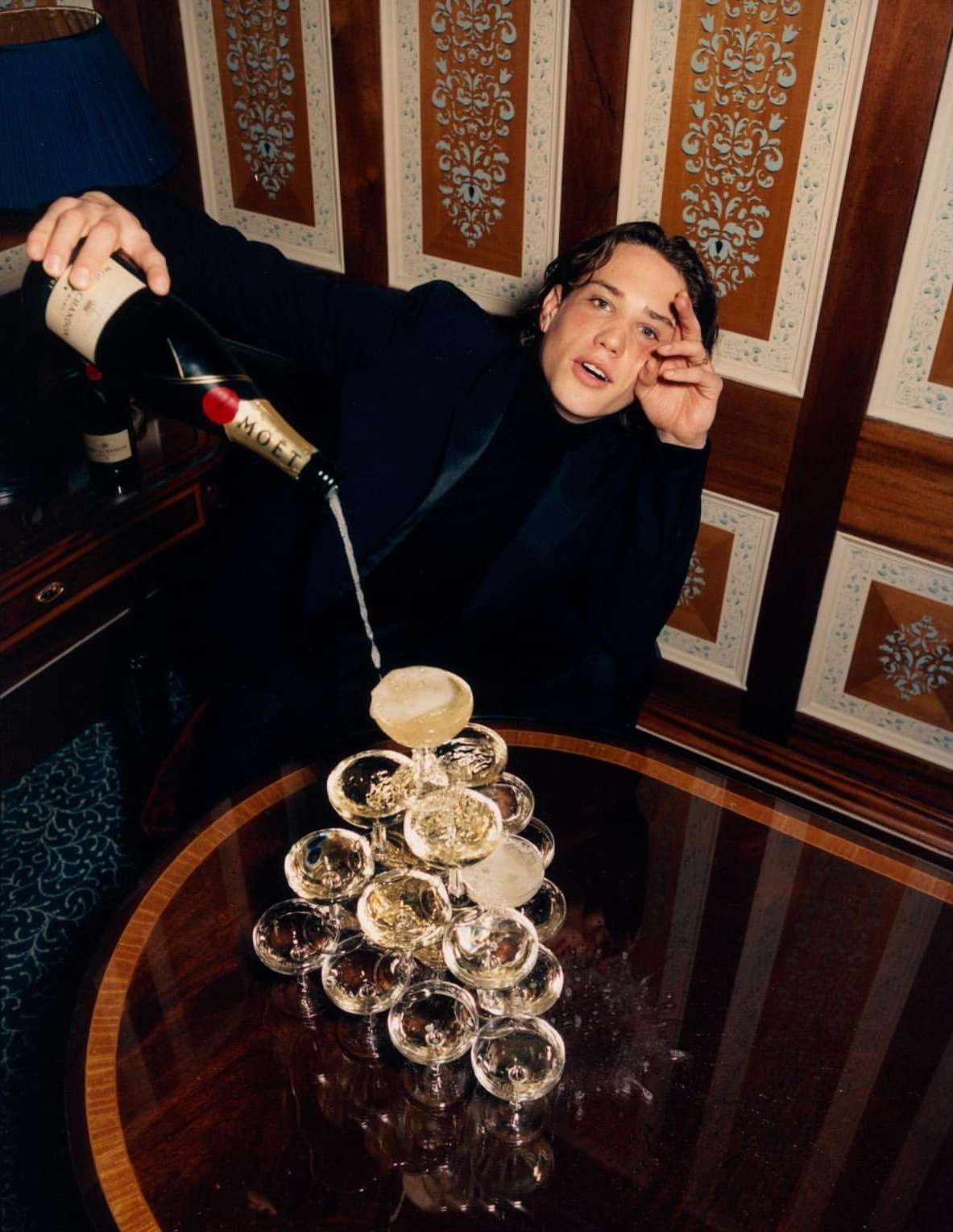 Adult man in black suit pours champagne into tower of glasses indoors near patterned wall, dark wood furniture, bottle of Moët.