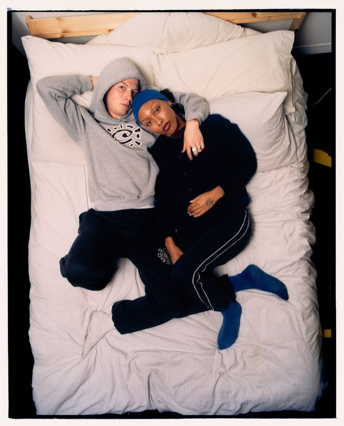 Overhead shot of a couple lying on a striped mattress in casual attire. The man is in a hoodie, and the woman wears a blue headscarf.