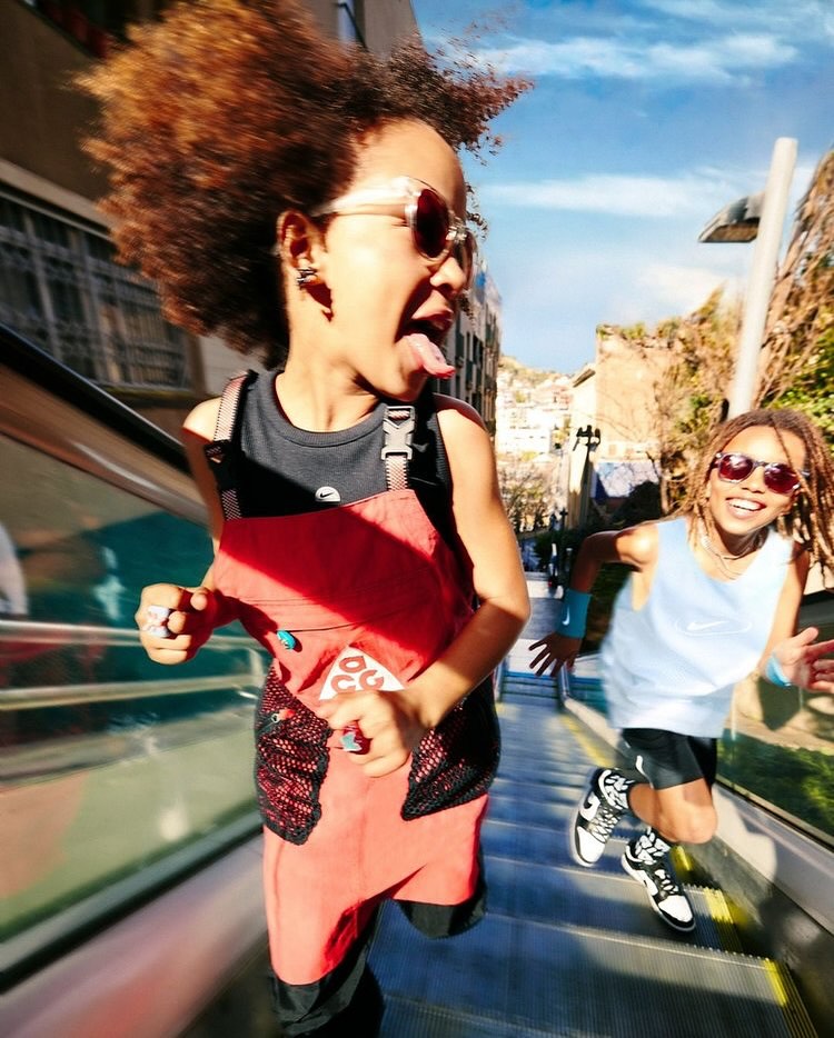 Two children run down a metal escalator outdoors on a sunny day, one sticks out their tongue, both wear sunglasses. Buildings visible.