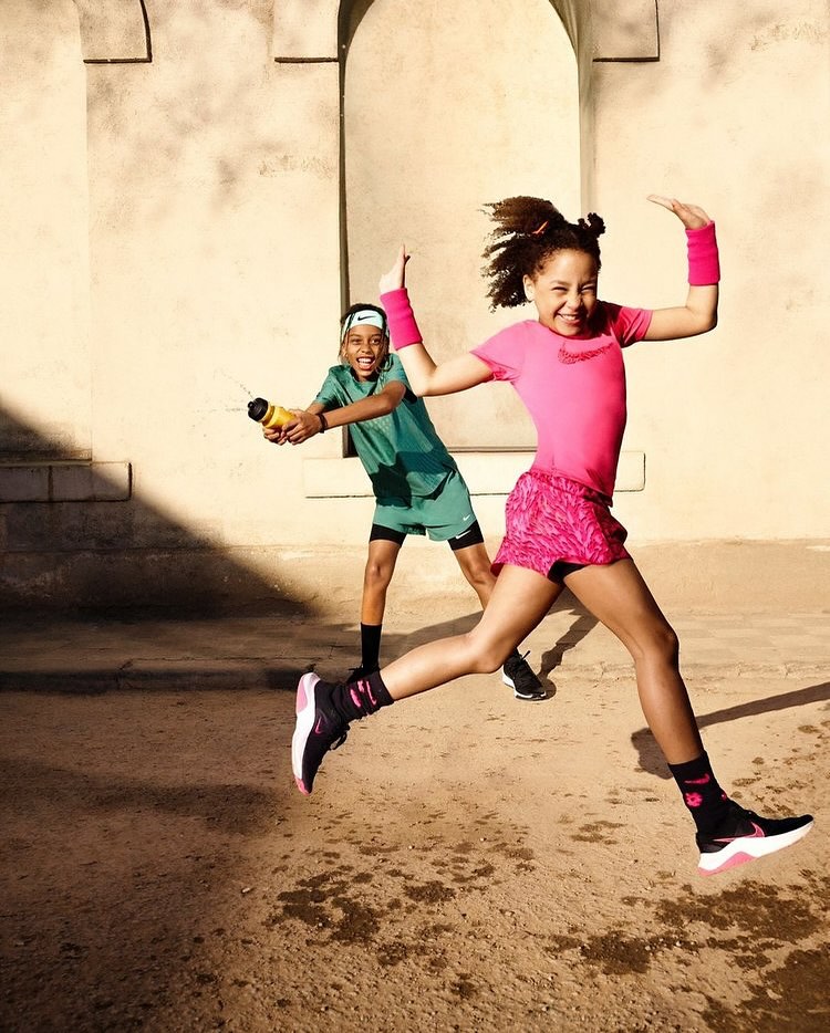Two children jumping in the air with water bottle in an urban setting. One child wears pink outfit, other child wears green athletic wear.