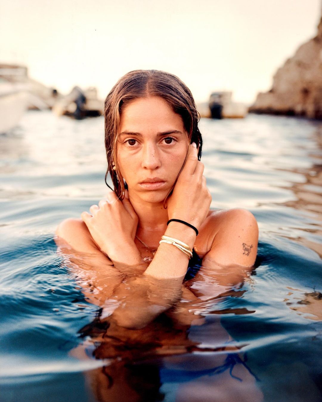 Portrait of a woman in water, with her hands touching her face, boats and land in the background, outdoor setting, natural light.