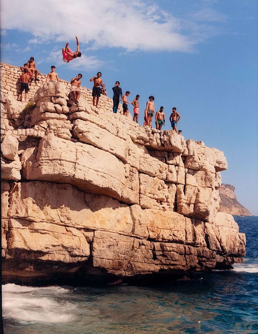 A group of young men and boys are on a stone cliff on the beach, with one jumping in mid-air against blue sky with scattered clouds.