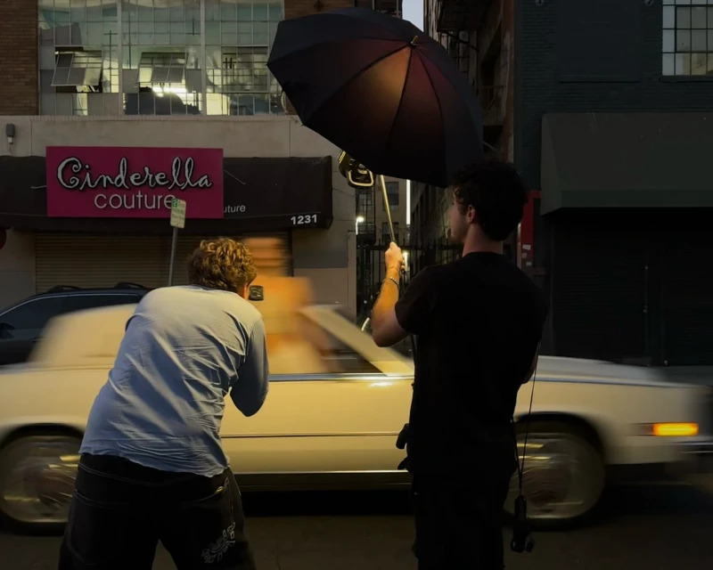 Street scene at dusk with two adult males; one holding an umbrella, the other using a camera, a blurred car, and Cinderella Couture sign.
