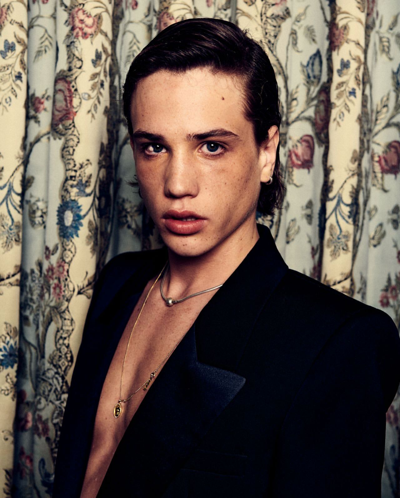 Portrait of a young adult male in a black suit against a floral curtain backdrop, showcasing fashion and editorial style.