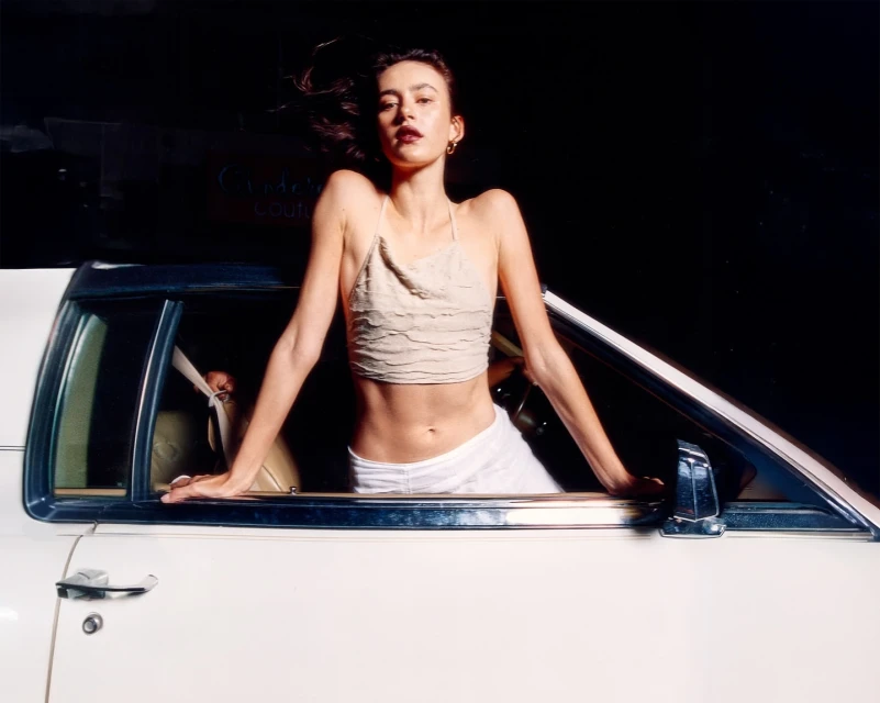 Woman in beige top leans out of a white car with a black interior. Dark background with sign, outdoor, medium shot, portrait orientation.