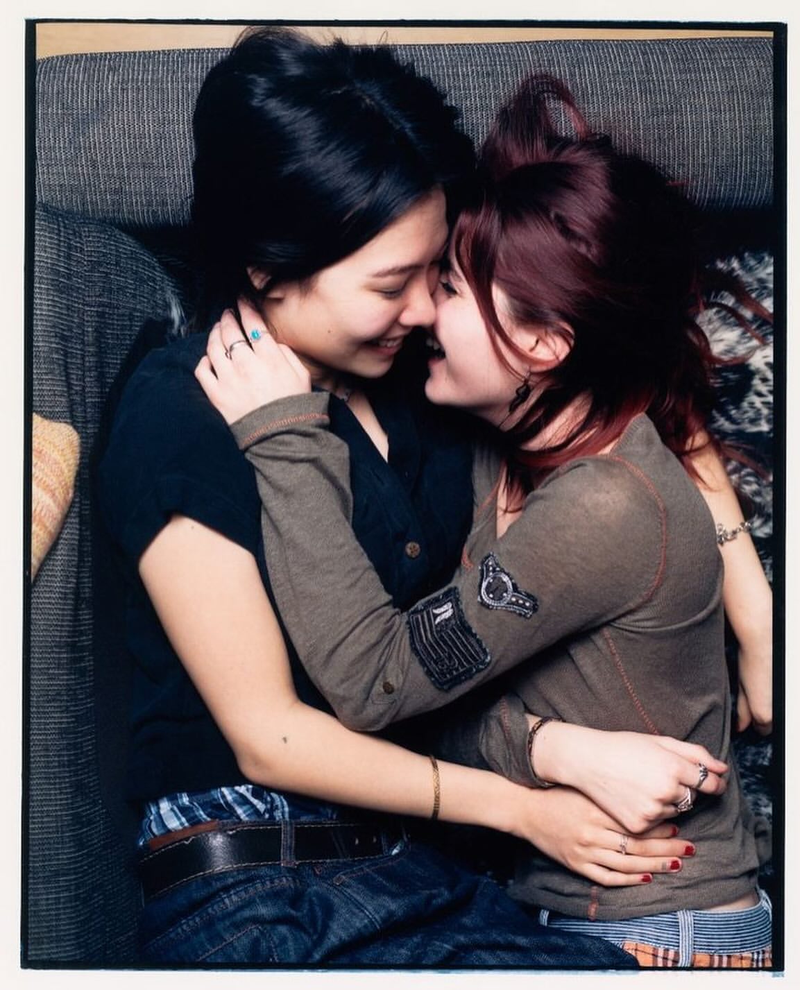 Two young women embracing indoors. One woman with dark hair in a black top; the other has auburn hair, wearing a grey long-sleeved shirt.