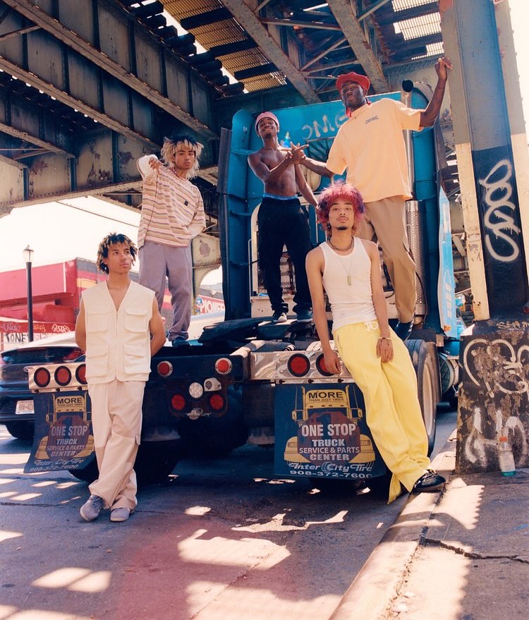 Group of five stylish young adults posing in front of a semi-truck under a bridge. Urban setting with neutral, yellow, and blue tones.