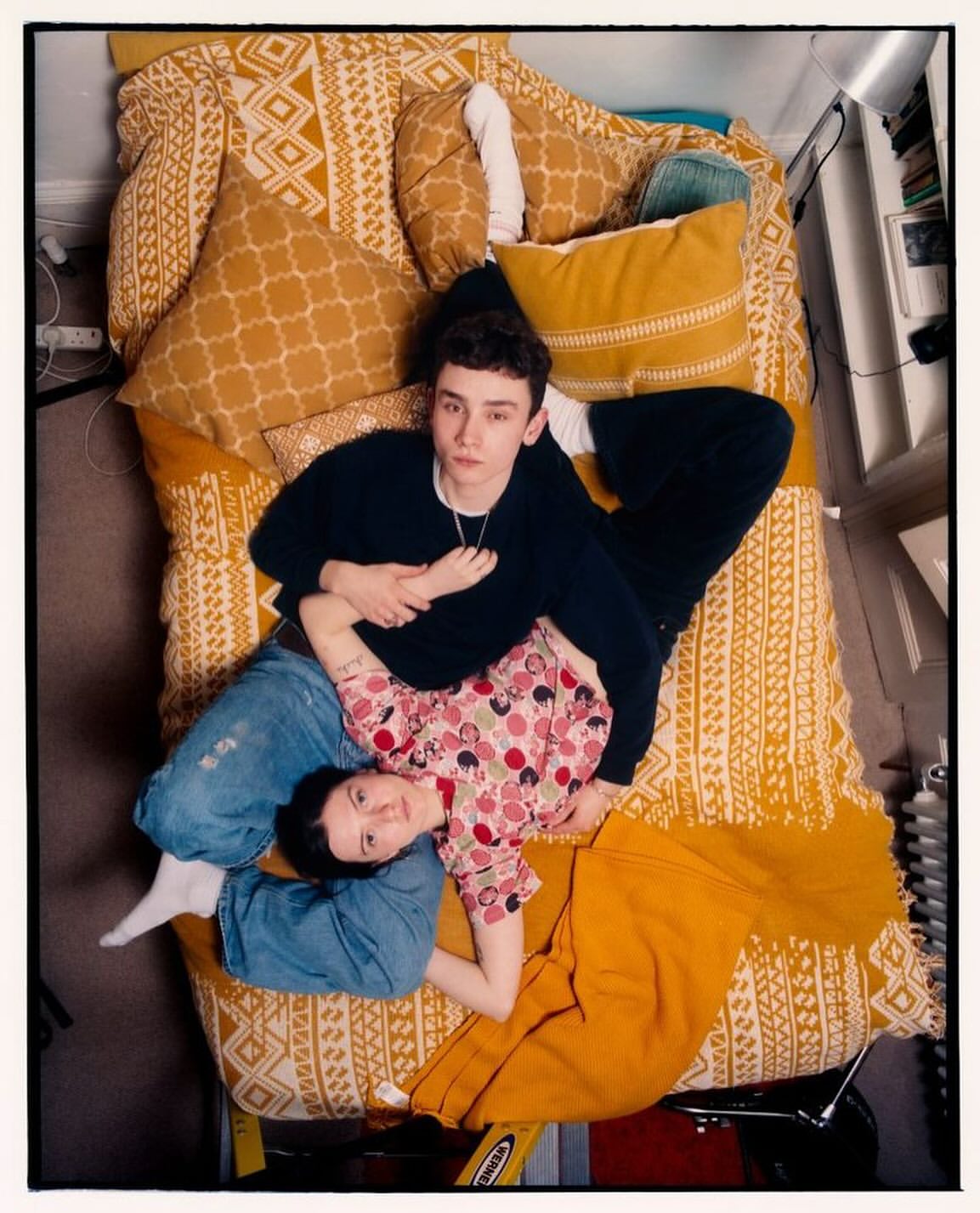 Overhead shot of a couple lying on a mustard-yellow patterned bed with pillows. The woman is wearing a patterned top and jeans.