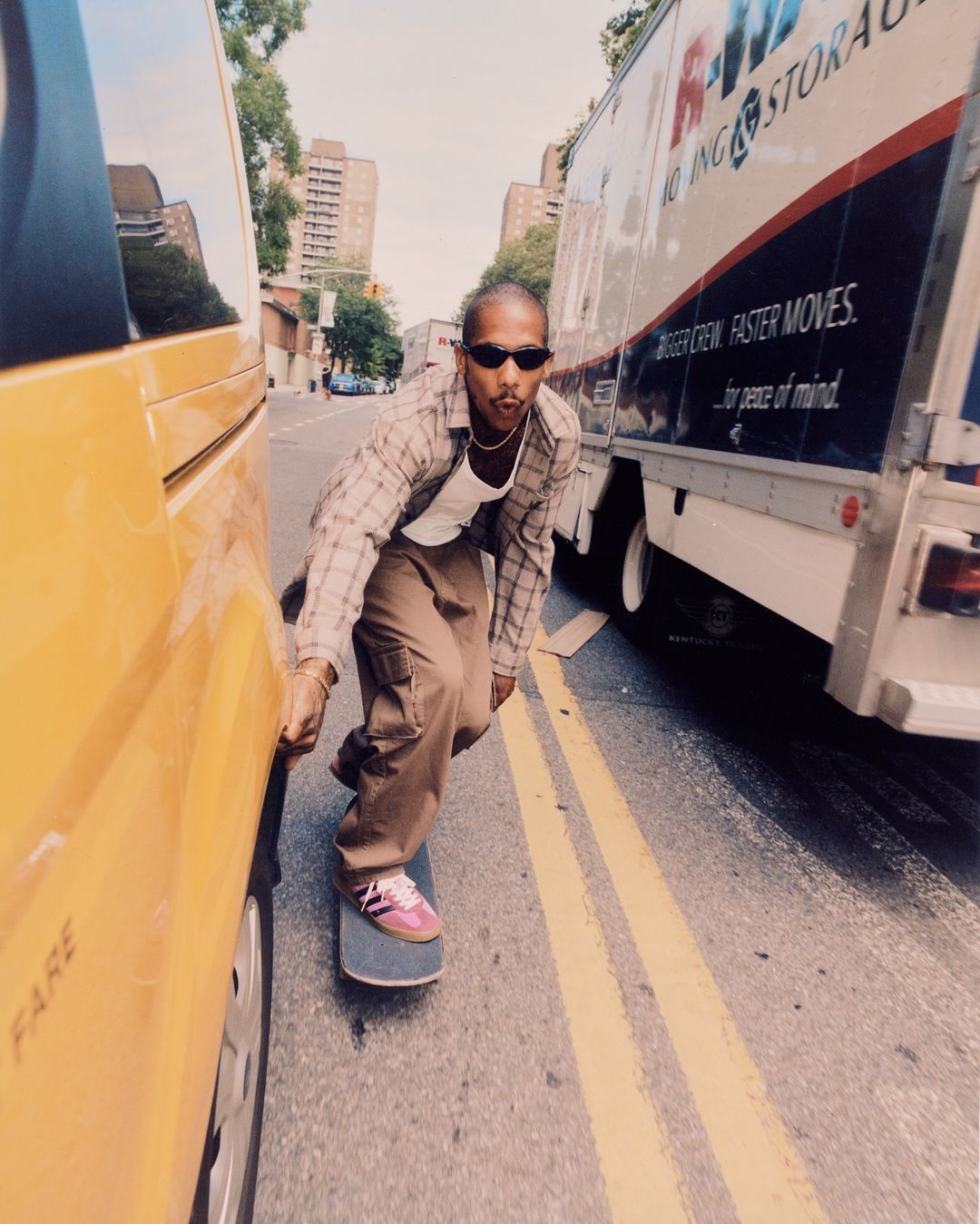 Man skateboarding in plaid shirt, cargo pants, sunglasses on city street between yellow cab and moving truck, captured in bright daylight.