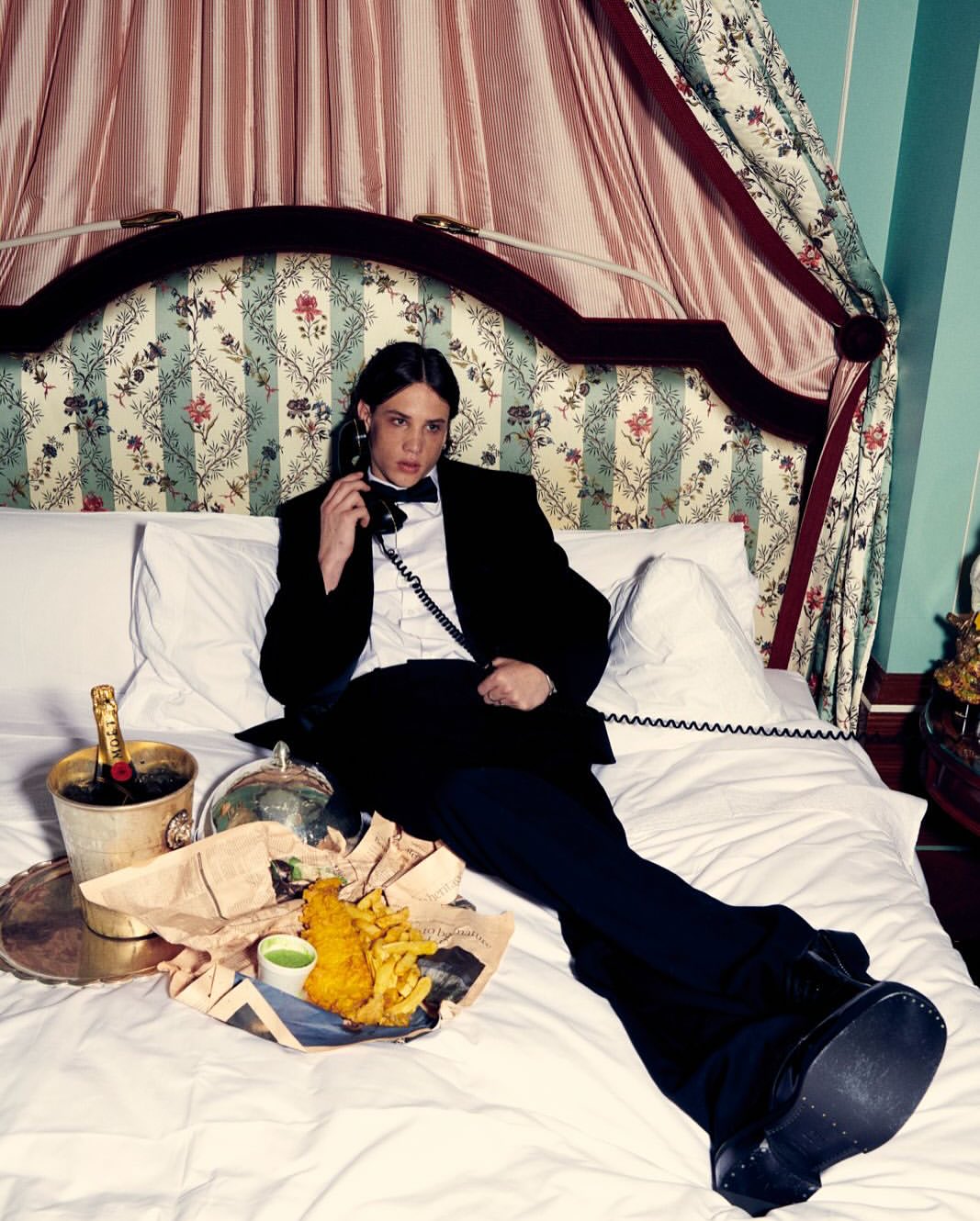 Man in a tuxedo lounging on a bed with fish and chips and Moët & Chandon while talking on the phone in an indoor setting.