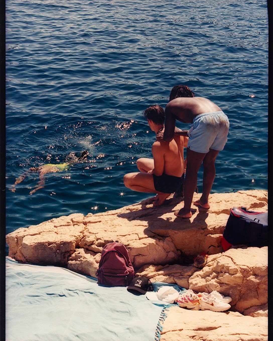 Two young men relax on a rocky outcrop overlooking the blue sea as a person swims in the distance on a sunny day at the coast.