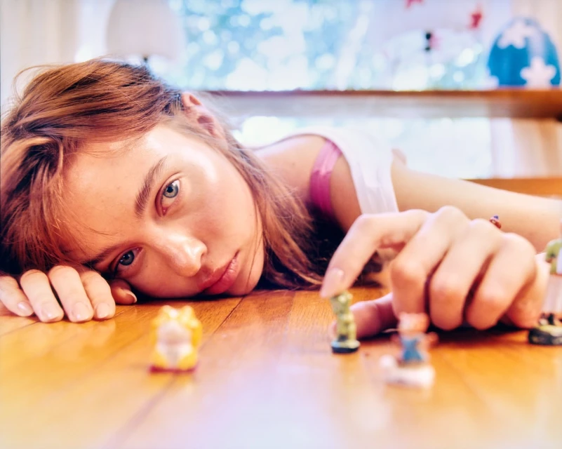 Woman lying on a wooden surface, with her hand reaching towards miniature animal figures, bathed in soft natural light.