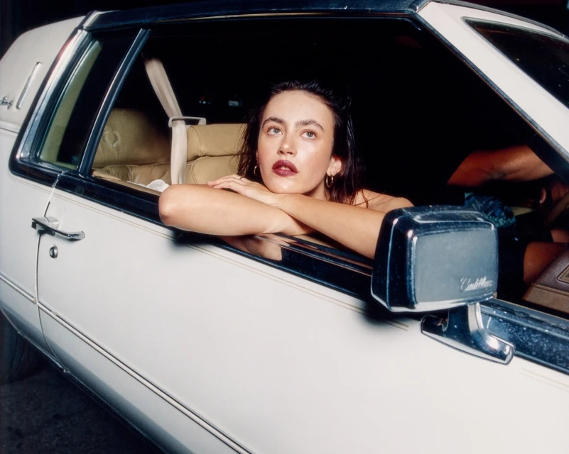 Young woman leans on car window in evening light, dark lipstick, tan interior, vintage vehicle, posing inside a classic car.