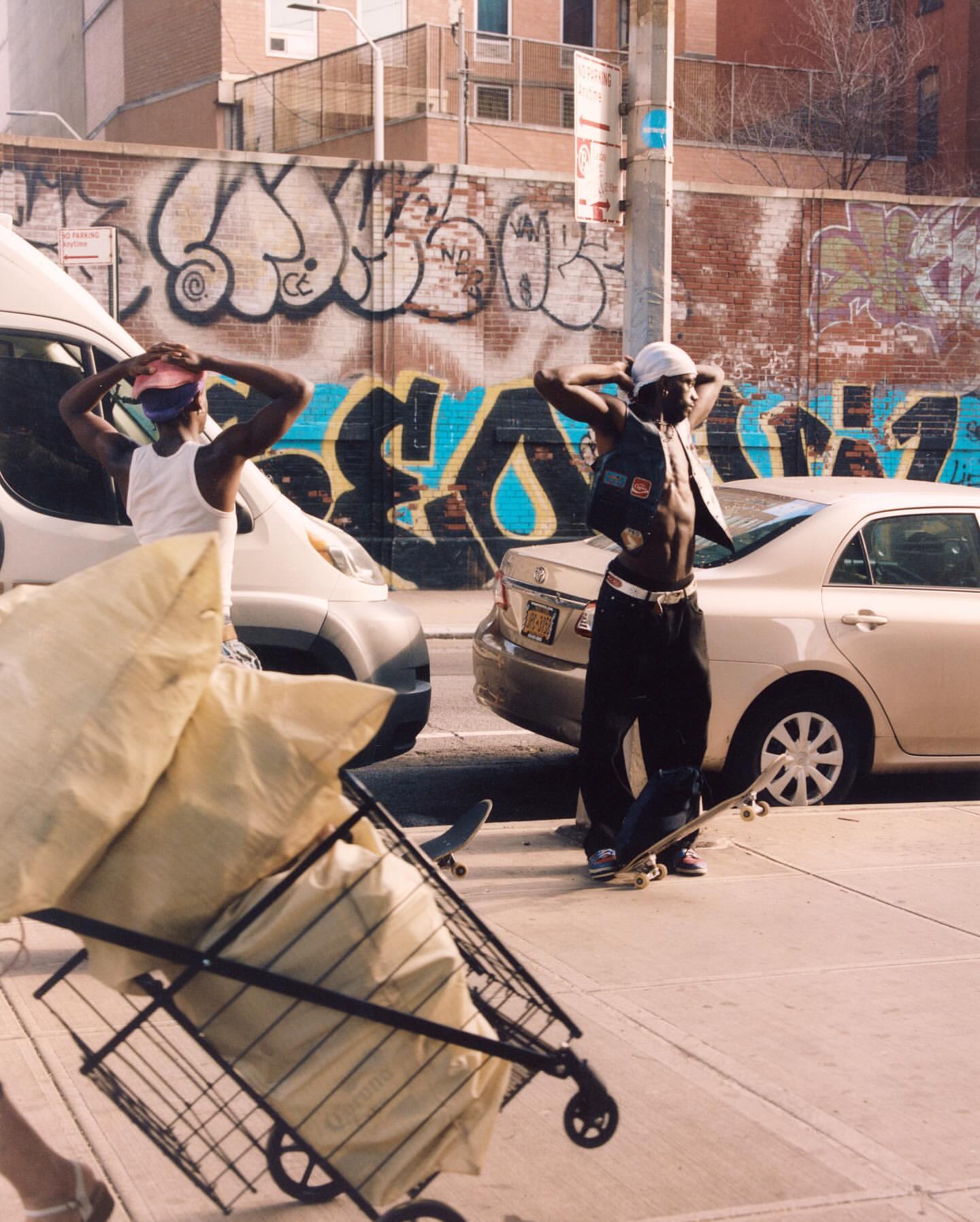 Two African American men in street clothing stand near cars and a shopping cart loaded with bags, with a graffiti wall in the background.