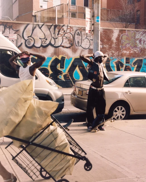 Two African American men in street clothing stand near cars and a shopping cart loaded with bags, with a graffiti wall in the background.