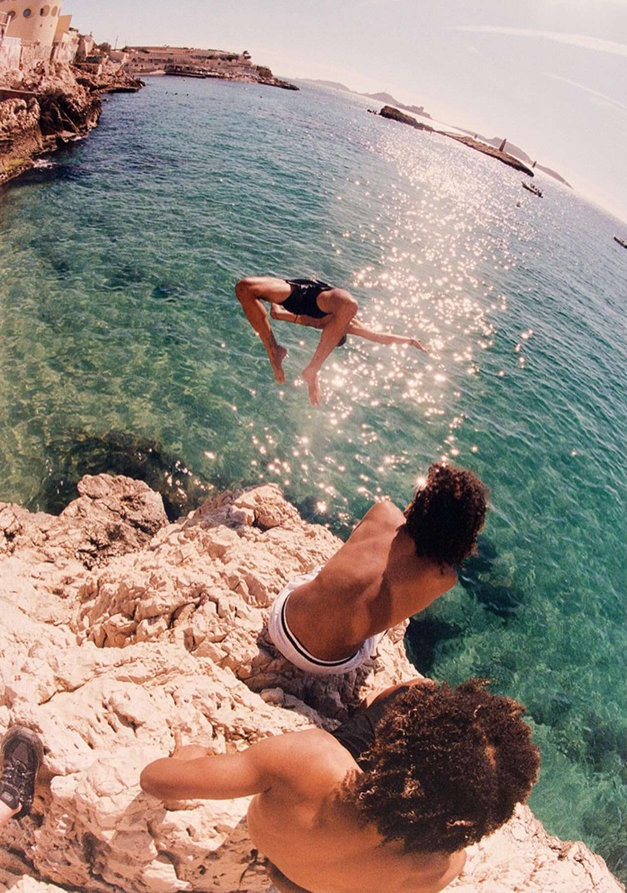 A man in mid-air performing a flip over crystal-clear ocean water, captured with a wide-angle lens showing the rocky coastline.