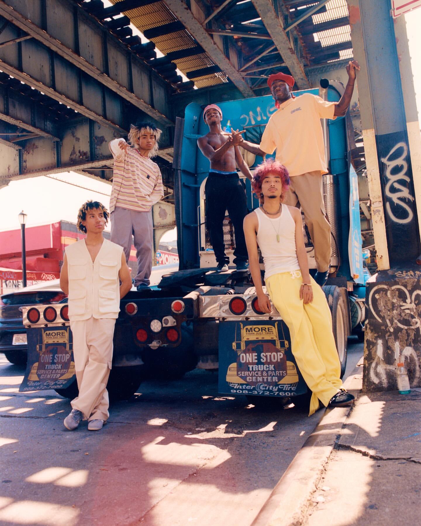 Five young adult models strike fashion poses on the back of a semi-truck under a bridge on a city street in the daytime in bright sunlight.